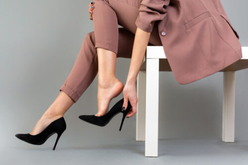 The concept of varicose disease. A woman in a suit takes off her high-heeled shoes. Legs close up. Gray background.